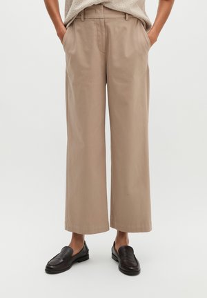 Person trägt beige weit geschnittene kurze Hosen, schwarze Loafers und ein beiges, strukturiertes Oberteil mit Händen in den Taschen.