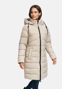 Vrouw in een beige gewatteerde lange winterjas met capuchon en zwarte rits, staande met ontspannen handen langs haar zij, kijkend opzij.