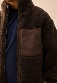 Brun fleece zip-up jakke med høj krave, der har en tekstureret overflade og en stor mørk stoflomme på venstre side.