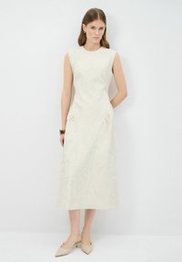 Mouwloze crèmekleurige jurk met textuurpatronen van bladeren; knielange silhouet met zijzakken en subtiele draping; gecombineerd met beige hoge hakken.