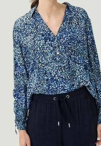 Blouse à motifs bleus et verts avec un col, une patte de boutonnage et des manches longues, associée à un pantalon élastique bleu marine.