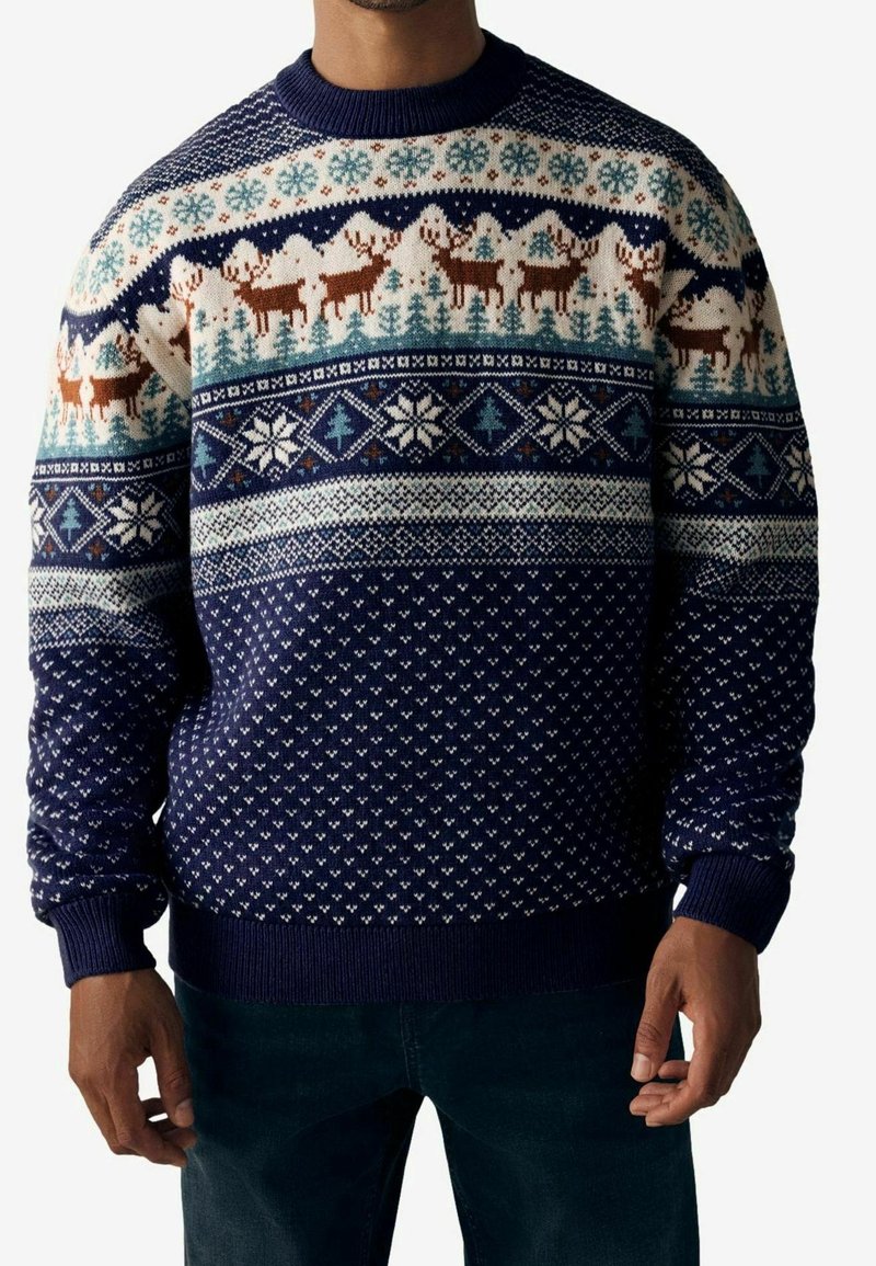 Homme portant un pull d'hiver bleu marine avec des motifs de rennes, flocons de neige et arbres en blanc, marron et bleu clair.