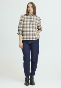 Beige-und-braun-checkierte Bluse mit Kragen und Puffärmeln, kombiniert mit dunkelblauen, taillierten Jeans und schwarzen Knöchelstiefeln.