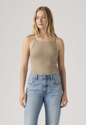 Lyshåret kvinde med langt hår iført en beige ærmeløs top og højttaljede lyseblå jeans, stående mod en ensfarvet baggrund.