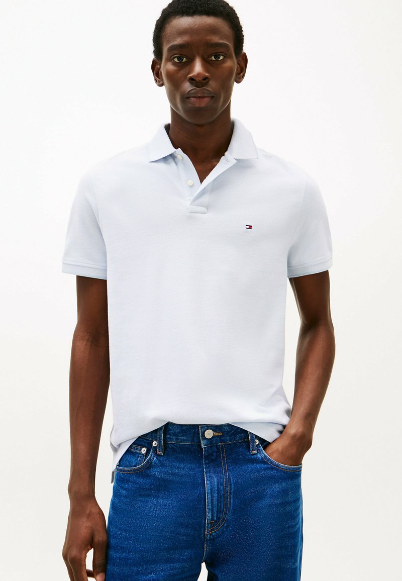 Polo shirt azzurro chiaro con maniche corte, in cotone, e un piccolo logo. Indossato con jeans blu scuro. Colletto semplice e dettaglio dei bottoni.