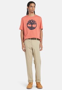 T-shirt corallo a maniche corte con logo di un albero blu navy, pantaloni khaki e stivali marroni. Presenta una vestibilità comoda e una texture liscia.