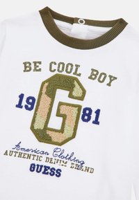 T-shirt en coton blanc avec col côtelé vert olive ; présente un texte brodé en brun, bleu et camouflage sur le devant, y compris un grand "G".