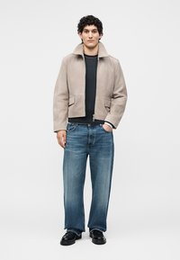 Junger Mann mit lockigem Haar, der eine beige Jacke mit Reißverschluss, ein schwarzes Shirt, blaue Jeans und schwarze Schuhe trägt und mit einer Hand in der Tasche steht.