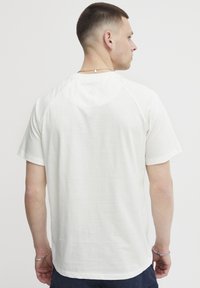 Hvit kortermet t-skjorte laget av myk stoff, med raglan-design og en subtil preget logo på øvre rygg.
