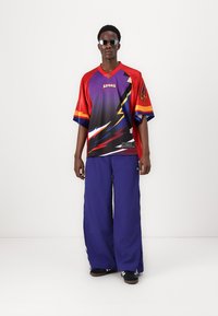 Red and purple short-sleeve jersey with a lightning graphic; paired with loose-fitting blue pants. Features sunglasses and black sneakers.