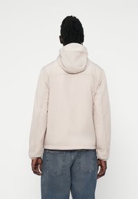 Šviesiai rožinė su kapu raišteliu striukė su lygiu paviršiumi, laisvas kirpimas ir elastingos rankovės, derinama su mėlynomis džinsinėmis kelnėmis.