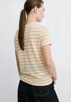 Kobieta z brązowym kucykiem, ubrana w żółtą koszulkę w paski z krótkim rękawem i ciemne dżinsy, stojąca bokiem na jednolitym tle.