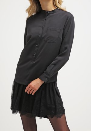 Vrouw draagt een zwarte blouse met lange mouwen, knopen en een borstzakje en een zwarte kanten rok tegen een effen achtergrond.