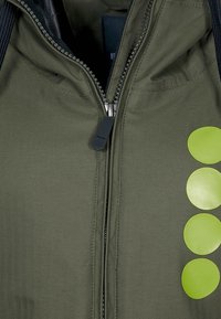 Giacca verde oliva con un motivo herringbone testurizzato, dotata di una chiusura centrale con zip e quattro evidenti accenti rotondi di colore verde lime.