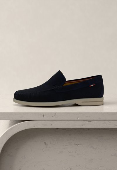 Μπλε σουέντ μοκασίνι με λευκή σόλα, ραφές μοκασίνου και μικρή υφασμάτινη λεπτομέρεια κόκκινη, λευκή και μπλε στο πλάι, εκτεθειμένο σε λευκή επιφάνεια.