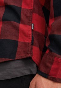 Camicia flanella a quadri rossa e nera con cuciture visibili, etichetta di lato e indossata sopra una maglietta grigia. Texture morbida e vestibilità comoda.