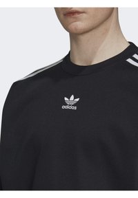 Musta huppari, jossa on pyöreä pääntie, jossa on valkoinen Adidas-logo ja kolme valkoista raitaa olkapäillä. Silkkinen, tekstuuriaines.