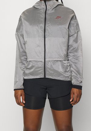 Silver vindjacka med svart rutmönster, hel dragkedja, justerbar huva och fickor, i kombination med svarta shorts. Lätt och strukturerad tyg.