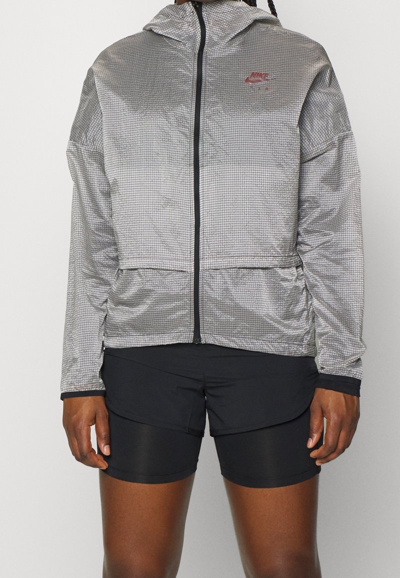 Stříbrná větrovka s černou mřížkovanou vzorem, plným zipem, nastavitelnou kapucí a kapsami, v kombinaci s černými šortkami. Lehká a strukturovaná tkanina.