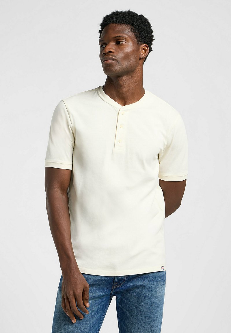 Kortærmet henley skjorte i lys creme med knaplukning og rund halsudskæring, lavet af glat materiale, parret med blå jeans.