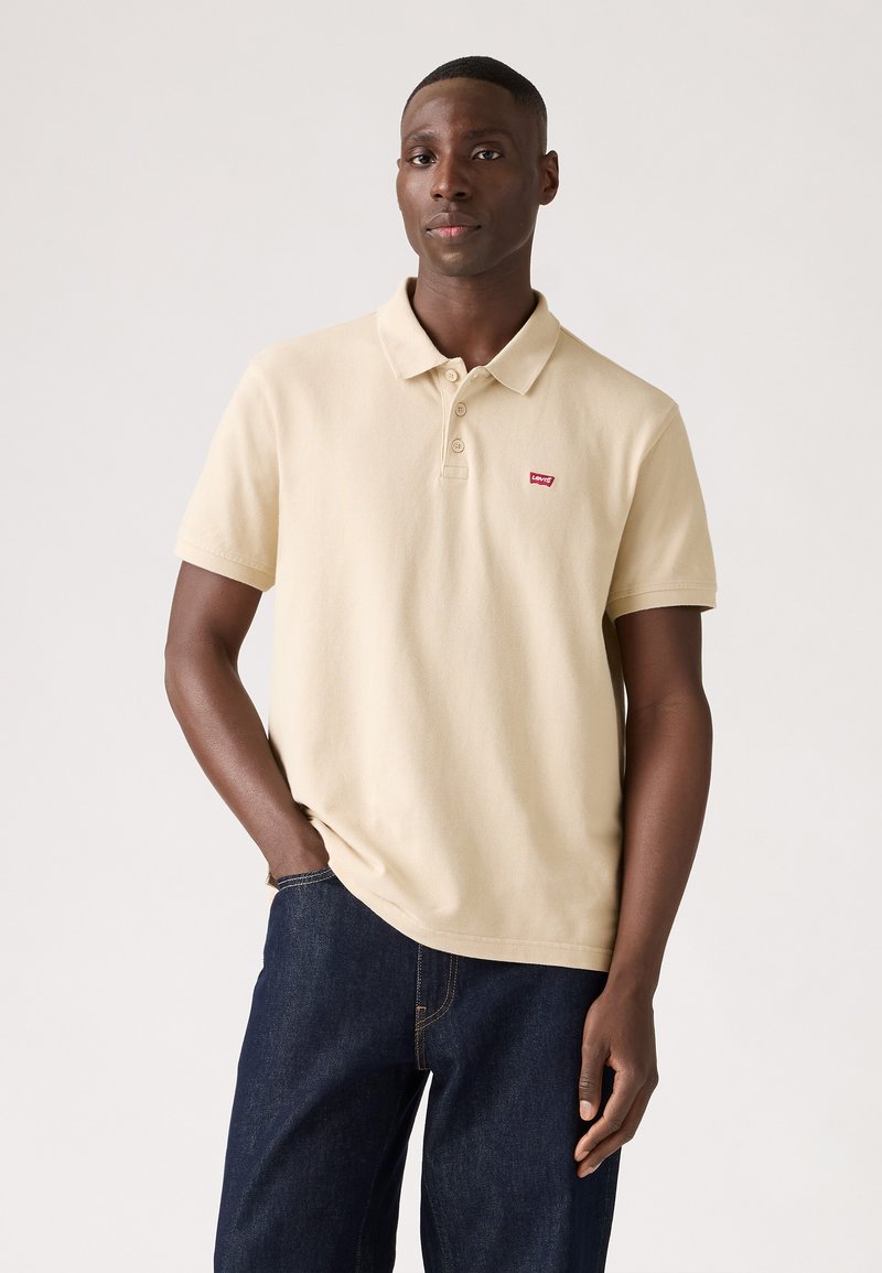 Camisa polo beige de algodón, con cuello, tapeta de tres botones y un pequeño logotipo rojo en el pecho. Combinada con jeans oscuros.