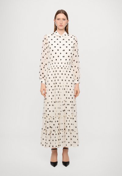 Lang, beige kjole med polka prikker, kraveudskæring, lange gennemsigtige ærmer og tiered ruffle kant. Båret med spidse sorte hæle.