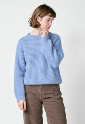Femme aux cheveux courts et bruns portant un pull tricoté bleu clair et un pantalon en velours côtelé marron, regardant vers le bas et touchant son menton.