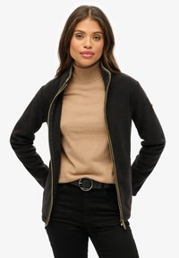 Czarna kurtka zapinana na zamek z wysokim kołnierzem i złotym zamkiem, noszona na beżowym golfie. Modelka ma również na sobie czarne spodnie i okrągły pasek.