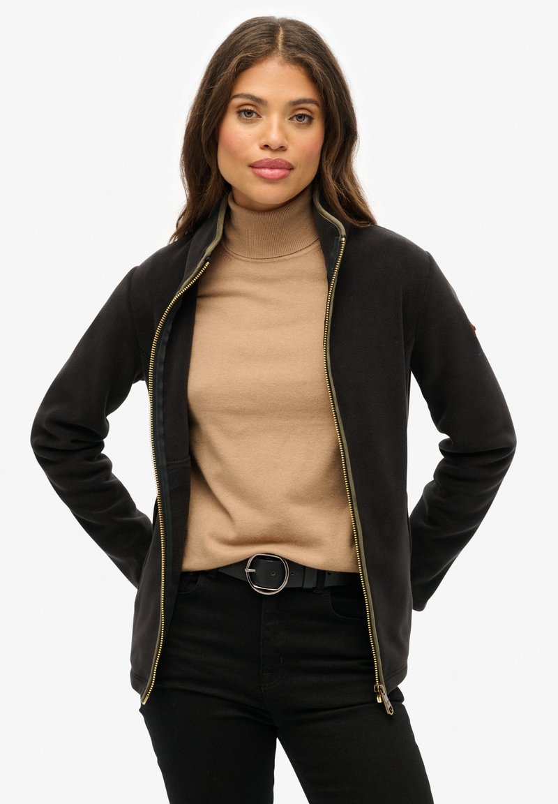 Czarna kurtka zapinana na zamek z wysokim kołnierzem i złotym zamkiem, noszona na beżowym golfie. Modelka ma również na sobie czarne spodnie i okrągły pasek.