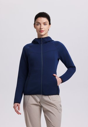 Frau trägt eine marineblaue Kapuzenjacke mit Reißverschluss und beige Hose, steht mit einer Hand in der Tasche vor einem schlichten Hintergrund.