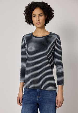 Kobieta z krótkimi kręconymi włosami, ubrana w granatowo-biały pasiasty długi rękaw i niebieskie dżinsy, stojąca na neutralnym tle.