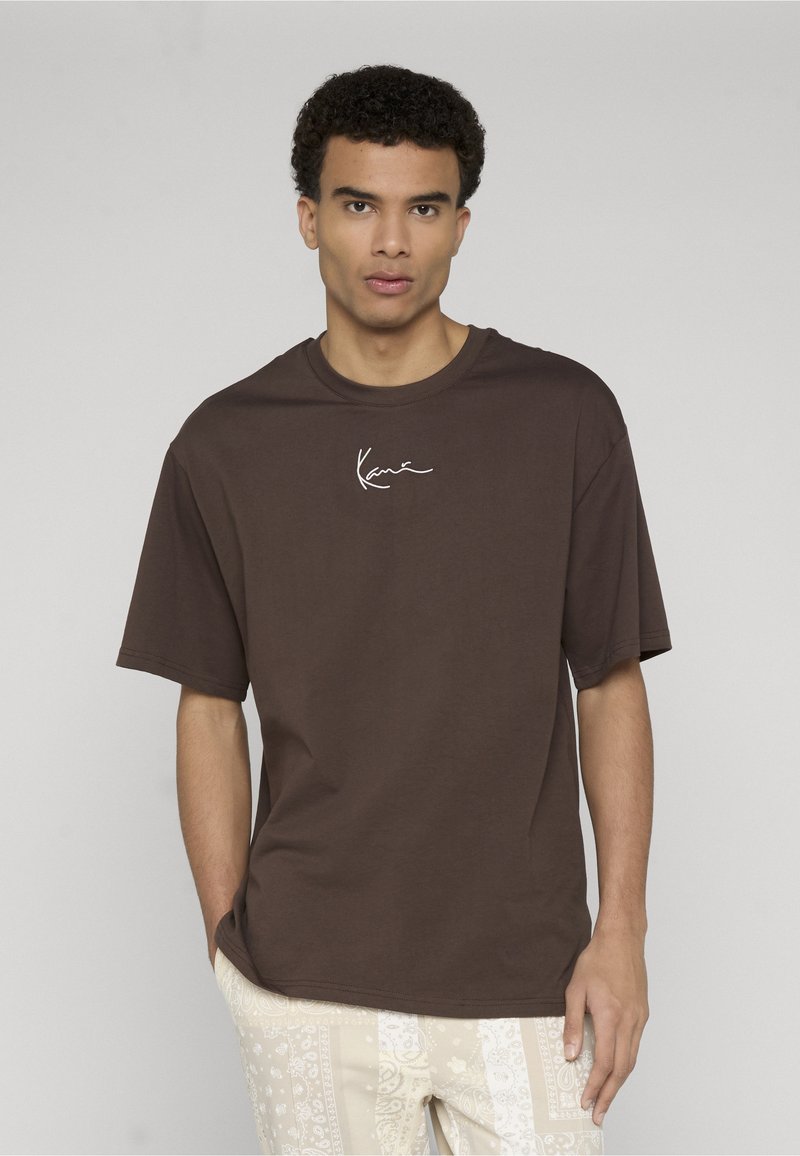 Fiatal férfi rövid göndör hajjal, barna pólót visel fehér aláírásos szöveggel és világos mintás nadrágot, zsebre tett kézzel áll.