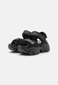 Sandales noires avec des sangles en tissu texturé et des fermetures Velcro réglables. Semelle en caoutchouc épaisse avec des rainures prononcées et des détails de logo.