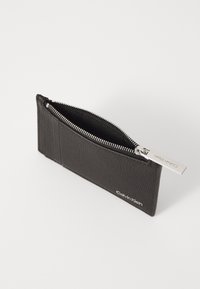 Zwarte leren ritstas met een gestructureerd oppervlak, voorzien van een zilveren rits en een Calvin Klein-logo dat aan de zijkant is gestempeld.