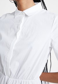 Camicia bianca con colletto a bottoni e vita arricciata. Tessuto liscio con un aspetto leggermente stropicciato.