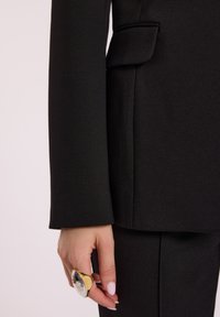 Dłoń z jasnoróżowymi paznokciami, nosząca dużą złotą i srebrną pierścionek, spoczywająca obok kieszeni czarnej marynarki i spodni.