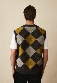 Ärmellose Strickweste in Dunkelbraun mit grauem und senffarbenem Rautenmuster, mit weicher Textur und rundem Ausschnitt, über einem weißen Hemd getragen.