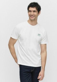 Hvid bomulds t-shirt med korte ærmer, rund halsudskæring og grøn tekst "1974" på venstre bryst, sat sammen med mørk denim jeans.
