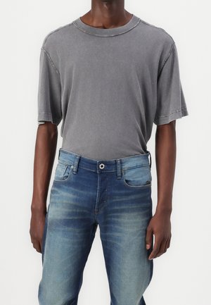 Homme portant un t-shirt ample gris délavé rentré dans un jean bleu délavé avec poches avant et fermeture par boutons.