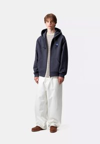 Felpa con zip grigio scuro con un logo blu, indossata sopra una maglietta a righe grigio chiaro, abbinata a pantaloni bianchi larghi e scarpe marroni.