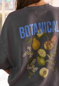 Felpa grigia scura con una grande stampa botanica di frutti e fiori sul retro, con la parola "BOTANICAL" in grassetto blu.