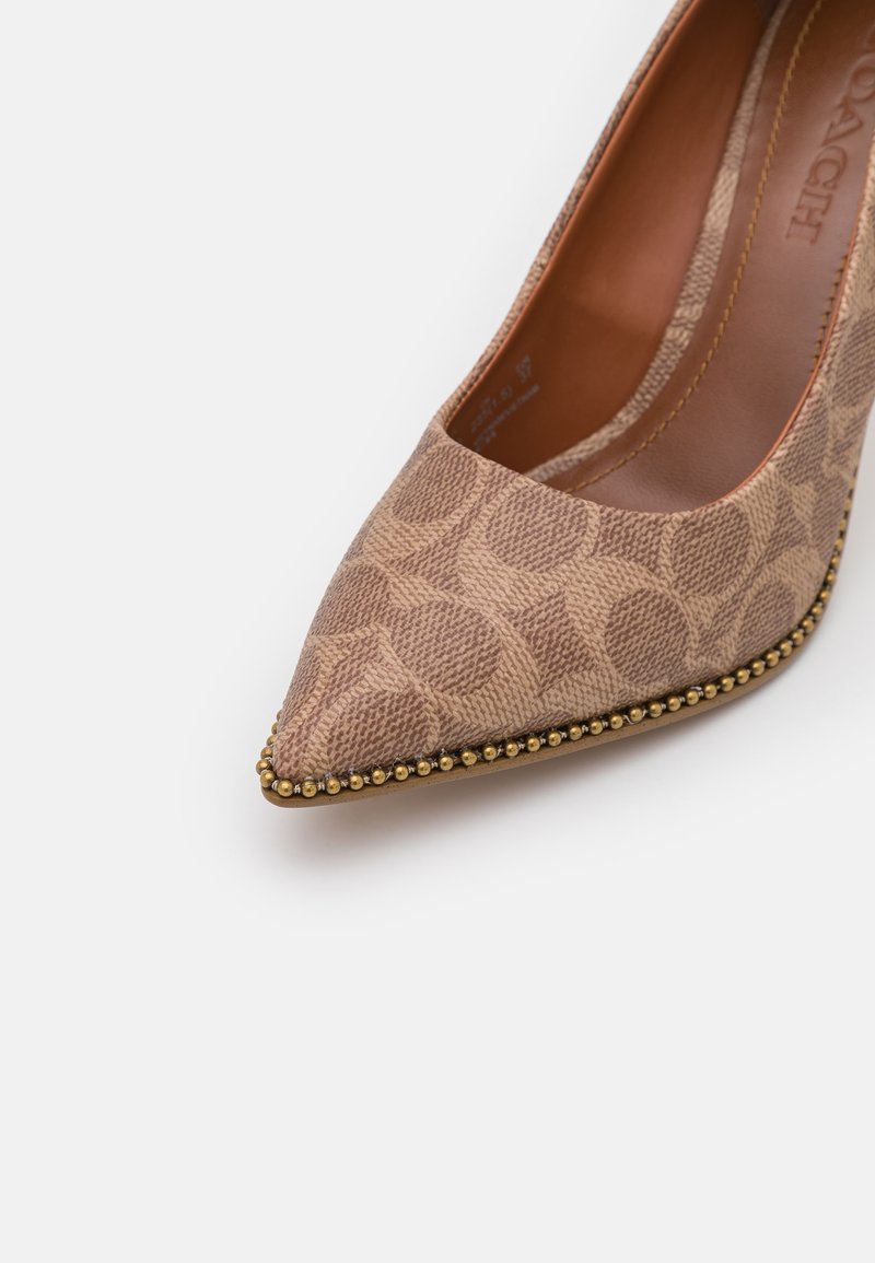 Zapato de tacón beige con punta afilada y un patrón texturizado, adornado con tachuelas en tono dorado a lo largo de la suela. Interior suave y detalles de costura sutiles.