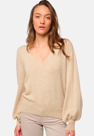 Pull en tricot beige avec décolleté en V, manches ballon et poignets côtelés. Texture douce avec une coupe ample, associé à un pantalon de couleur claire.