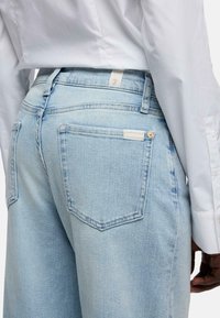 Jeans in denim azzurro chiaro a vita alta, con una tasca posteriore decorata con un'etichetta e un rivetto dorato, abbinati a una camicia bianca.