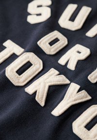 Sweatshirt en coton avec broderie blanche en relief écrivant "SUPER TOKYO" sur un fond bleu marine foncé. Texture douce avec une finition lisse.