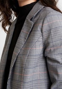 Blazer à carreaux gris avec des accents bleus et oranges, doté d'un revers structuré et d'un tissu texturé. Porté sur un col roulé noir.