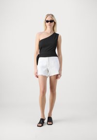 Svart top med én skulder og tekstur, kombinert med hvite shorts med frans og åpne svarte hæler. Modellen har også på seg mørke solbriller.