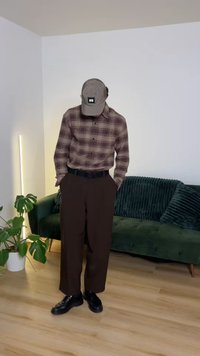 Brązowa koszula w kratę z guzikami, ciemnobrązowe spodnie, czarny pasek, czarne buty oraz szara czapka z naszywką. W tle zielona kanapa i roślina.