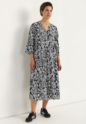 Frau in einem schwarz-weiß geblümten Midikleid mit 3/4-Ärmeln und schwarzen Turnschuhen, die vor einer weißen Wand steht.