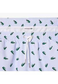 Costumi da bagno azzurri chiari con un motivo ripetitivo di coccodrilli verdi, vita elastica con cordino bianco e etichetta Lacoste.